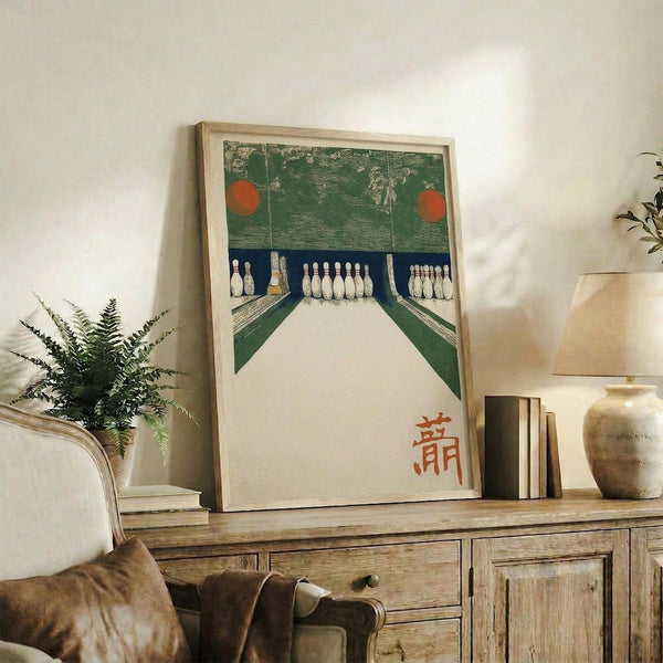 May include: Framed vintage-style bowling alley artwork displayed on rustic wooden sideboard in neutral living room. Print shows green textured bowling lanes with white pins and two red balls, featuring orange Japanese characters. Light wood frame sits among fern plant, ceramic vase, books and cream lamp against white wall.