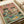 May include: Vintage-style poster print displayed on white paper showing two cats wearing sunglasses at a party table. One cat wears a colorful Hawaiian shirt, the other a striped shirt, both holding drinks. Table has tuna can, cheese, catnip fizz bottle, and small mice below. Pink background with multicolored "CAT PARTY" text at top. Cream ribbon visible in corner of photo.