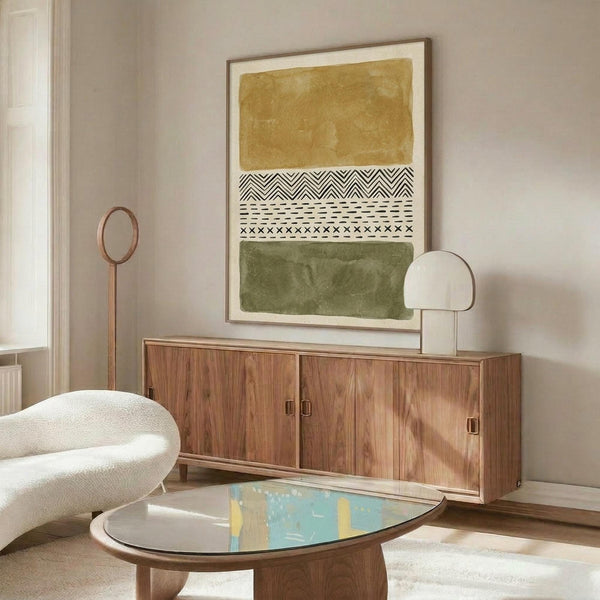 May include: Abstract art print with ochre yellow and olive green watercolor blocks separated by black and white tribal geometric patterns, displayed in thin wooden frame above mid-century walnut credenza. Modern living room setting with cream curved chair, oval glass coffee table, and contemporary table lamps on neutral walls.