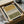 May include: Framed abstract art print with horizontal bands in ochre yellow and olive green separated by geometric tribal patterns in black and white. Print displayed in light wood frame, surrounded by brown cardboard packaging materials and wrapped wooden dowels on white tissue paper.