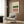 May include: Abstract geometric artwork with horizontal bands of green, white with dots, brown speckled section, and coral pink, displayed in light wood frame on wood-paneled wall above beige upholstered chair with geometric patterned pillow, brass floor lamp and dark round side table with open book visible in modern living room setting