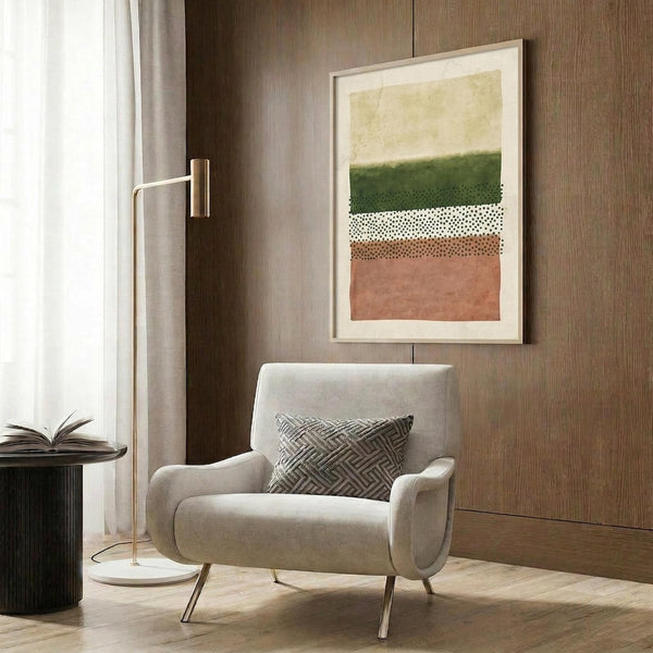 May include: Abstract geometric artwork with horizontal bands of green, white with dots, brown speckled section, and coral pink, displayed in light wood frame on wood-paneled wall above beige upholstered chair with geometric patterned pillow, brass floor lamp and dark round side table with open book visible in modern living room setting