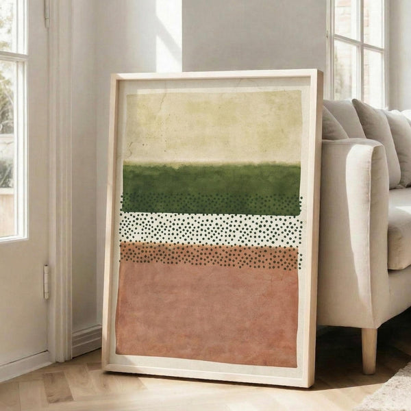 May include: Large framed abstract art print leaning against white wall next to beige sofa in bright living room. Artwork features horizontal color blocks in cream, forest green, coral pink with black dotted pattern band across middle. Light wood frame, natural lighting from windows, hardwood floors visible.