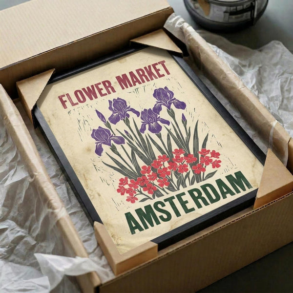 May include: Vintage-style poster print with cream background featuring purple iris flowers and red blooms, text reading "Flower Market" and "Amsterdam" in red and green lettering. Black wooden frame shown in cardboard shipping box with brown paper wrapping and tissue paper on white surface.