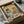 May include: Vintage-style poster print in black frame showing two illustrated cats at breakfast table - tabby cat in beret reading "Cat Tales" newspaper and orange cat in sunglasses and floral shirt holding coffee cup, with "CAT CAFE" text above, pastries and coffee items on checkered tablecloth, displayed in cardboard shipping box with protective wrapping on white surface