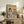 May include: Framed poster displaying two illustrated cats at a breakfast table - one tabby in black beret reading "Cat Tales" newspaper with coffee cup, another orange cat wearing sunglasses and tropical shirt. Table set with pastries, coffee pot, and checkered tablecloth. Vintage-style artwork with "CAT CAFÉ" text in blue and red letters against cream background. Frame sits on rustic wooden sideboard alongside books, ceramic vase, and fern plant in cozy living room setting.