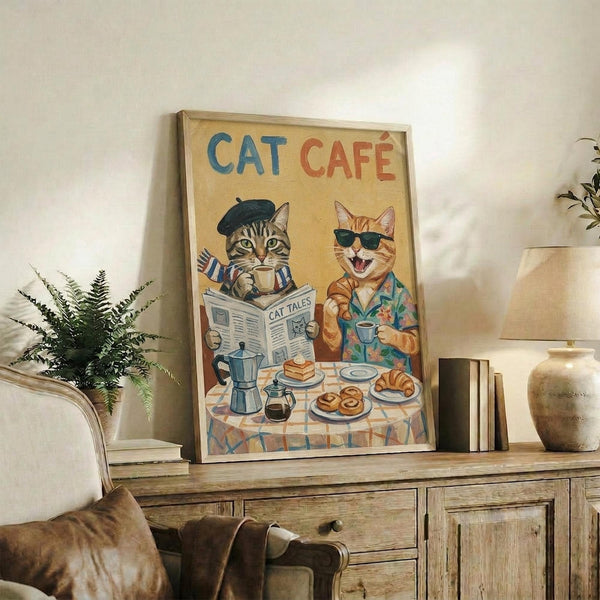May include: Framed poster displaying two illustrated cats at a breakfast table - one tabby in black beret reading "Cat Tales" newspaper with coffee cup, another orange cat wearing sunglasses and tropical shirt. Table set with pastries, coffee pot, and checkered tablecloth. Vintage-style artwork with "CAT CAFÉ" text in blue and red letters against cream background. Frame sits on rustic wooden sideboard alongside books, ceramic vase, and fern plant in cozy living room setting.