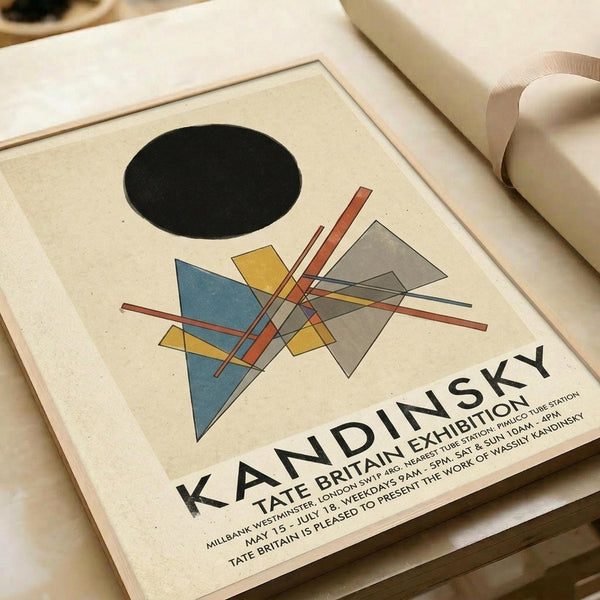 Abstract geometric composition featuring a black circle, intersecting colored shapes in blue, yellow, and gray, and crossing red and thin lines on cream background. Exhibition poster for Kandinsky show at Tate Britain, displayed flat on white surface with rolled paper nearby. Clean, minimalist presentation showcasing the modernist artwork and exhibition details in black text.