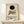 May include: Framed Kandinsky exhibition poster with abstract geometric design featuring black circle, colorful triangles, rectangles and intersecting lines in blue, yellow, red and gray. Light wood frame displayed on white wooden shelf beside small wrapped gift with dried plant sprig against cream wall.