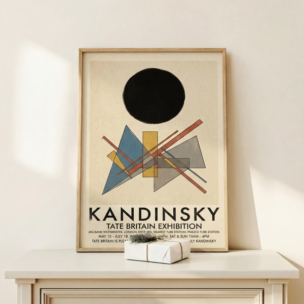 May include: Framed Kandinsky exhibition poster with abstract geometric design featuring black circle, colorful triangles, rectangles and intersecting lines in blue, yellow, red and gray. Light wood frame displayed on white wooden shelf beside small wrapped gift with dried plant sprig against cream wall.