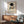 May include: Framed Kandinsky exhibition poster with abstract geometric shapes in blue, yellow, and red beneath a black circle, displayed above a wooden sideboard. Mid-century modern living room setting features a sage green upholstered armchair with wooden frame, small round coffee table, and white ceramic vase. Warm natural lighting creates shadows on white walls.
