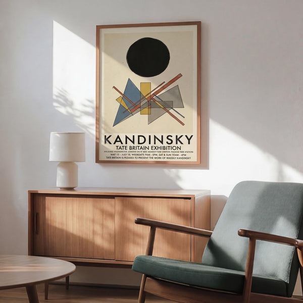 May include: Framed Kandinsky exhibition poster with abstract geometric shapes in blue, yellow, and red beneath a black circle, displayed above a wooden sideboard. Mid-century modern living room setting features a sage green upholstered armchair with wooden frame, small round coffee table, and white ceramic vase. Warm natural lighting creates shadows on white walls.