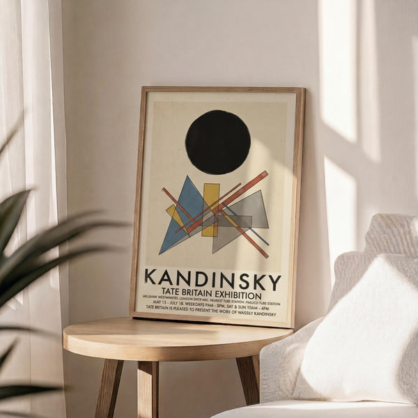 May include: A framed exhibition poster featuring abstract geometric shapes in blue, yellow, gray, and red lines beneath a large black circle, displayed on a light wood round side table next to a beige upholstered chair with a textured pillow. Soft natural light creates shadows across the neutral-toned room with white curtains and a plant visible in the corner.