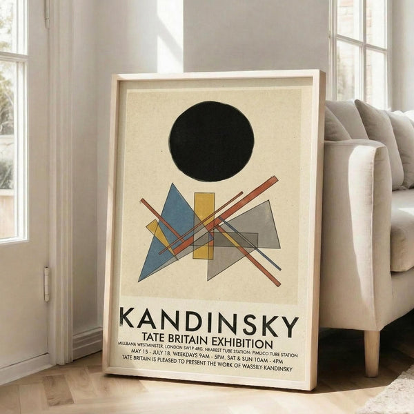 May include: Framed exhibition poster featuring abstract geometric composition with black circle, blue triangles, yellow rectangles, gray shapes, and red lines on cream background. Text reads "KANDINSKY TATE BRITAIN EXHIBITION" with venue details. Light wood frame leaning against white wall next to beige sofa in bright, minimalist living room with large windows and natural lighting.