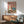 May include: Framed poster with pink background showing two cartoon ducks - one wearing chef's hat holding grilled fish, another in beanie and denim jacket looking puzzled. Kitchen scene with cutting board, vegetables, wine bottle, and cooked dishes scattered on counter. Displayed above mid-century modern wooden credenza with white ceramic lamp, next to sage green upholstered armchair in bright, naturally-lit living room with white walls.