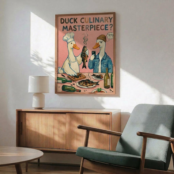 May include: Framed poster with pink background showing two cartoon ducks - one wearing chef's hat holding grilled fish, another in beanie and denim jacket looking puzzled. Kitchen scene with cutting board, vegetables, wine bottle, and cooked dishes scattered on counter. Displayed above mid-century modern wooden credenza with white ceramic lamp, next to sage green upholstered armchair in bright, naturally-lit living room with white walls.