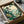 May include: Japanese woodblock print artwork in black frame being unboxed from cardboard packaging with tissue paper. The print shows a serene landscape with tall pine trees in foreground, rolling mountains in soft blues and greens, and small village buildings nestled in the valley below. The scene has muted earth tones with cream sky and depicts a peaceful countryside view in traditional ukiyo-e style.