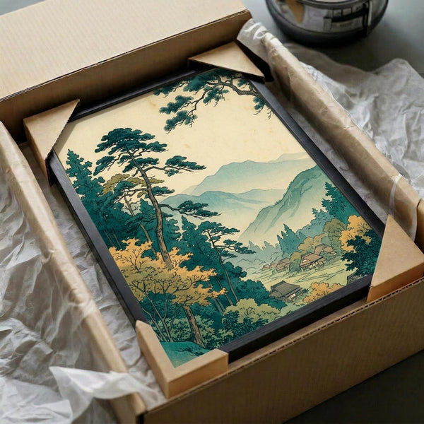 May include: Japanese woodblock print artwork in black frame being unboxed from cardboard packaging with tissue paper. The print shows a serene landscape with tall pine trees in foreground, rolling mountains in soft blues and greens, and small village buildings nestled in the valley below. The scene has muted earth tones with cream sky and depicts a peaceful countryside view in traditional ukiyo-e style.