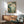 May include: Japanese woodblock print artwork in wooden frame showing mountain landscape with pine trees, traditional buildings, and misty blue-green hills. Displayed above mid-century modern walnut credenza with table lamp. Sage green upholstered armchair with wooden frame in foreground. Warm natural lighting creates shadows on white wall in minimalist living room setting.
