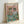 May include: Framed poster print leaning against white wall showing two anthropomorphic cats wearing sunglasses at a dining table. Left cat wears straw hat and floral shirt, right cat wears red bow tie and blue striped shirt. Pink background with green table displaying wine glasses, fish on plate, cat food bowls, tuna cans, and catnip brew bottle. Light wood frame with natural lighting creating soft shadows.