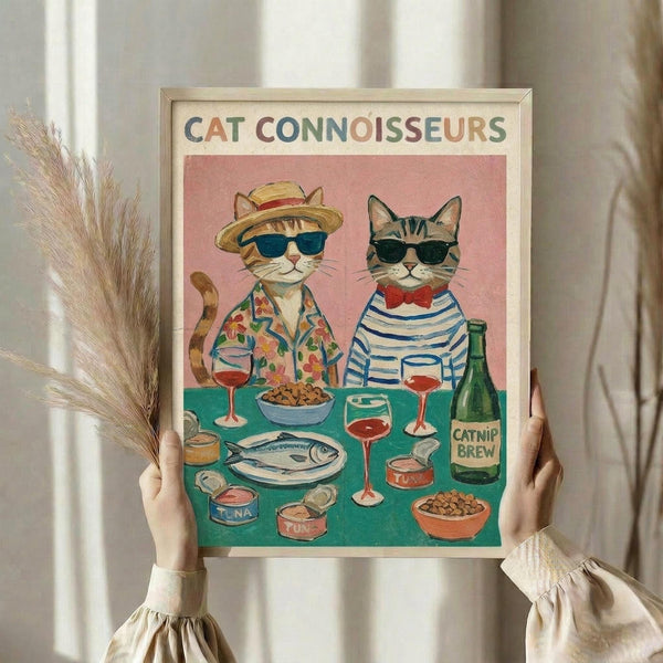 May include: Hands holding a framed poster showing two cats wearing sunglasses and clothing sitting at a turquoise table with wine glasses, fish, cat food bowls and canned tuna. The orange tabby cat wears a straw hat and floral shirt, while the gray striped cat sports a red bow tie and striped shirt. The poster has a pink background with "CAT CONNOISSEURS" text in colorful letters at the top. Natural wood frame against white curtains with dried pampas grass visible on the left side.