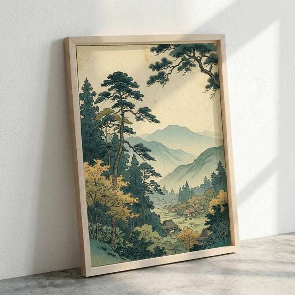 May include: Framed Japanese woodblock print artwork leaning against white wall on concrete floor. The vintage-style landscape shows tall pine trees in foreground with green and gold foliage, misty blue-green mountains in background, and small village buildings nestled in valley. Soft cream sky with muted color palette in traditional ukiyo-e art style, displayed in light wooden frame.