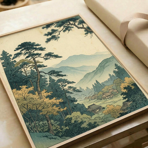 Japanese woodblock print showing a serene mountain landscape with tall pine trees in the foreground, rolling green hills, and traditional buildings nestled in a valley. The artwork features muted green, beige, and cream tones in classic ukiyo-e style. The print is displayed on cream paper within an open book or portfolio, photographed at an angle on a light wooden surface.