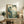 May include: Framed Japanese woodblock print artwork displayed on rustic wooden sideboard in neutral-toned living room. Print shows mountain landscape with tall pine trees, rolling hills, and traditional buildings in muted blue-green and cream tones. Scene includes fern plant in white pot, ceramic vase, stacked books, and brown leather chair. Natural lighting creates soft shadows on cream wall.