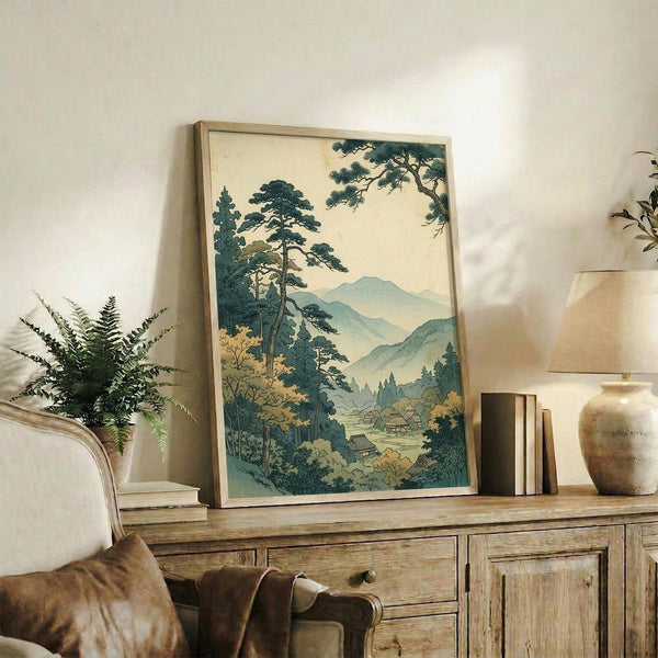 May include: Framed Japanese woodblock print artwork displayed on rustic wooden sideboard in neutral-toned living room. Print shows mountain landscape with tall pine trees, rolling hills, and traditional buildings in muted blue-green and cream tones. Scene includes fern plant in white pot, ceramic vase, stacked books, and brown leather chair. Natural lighting creates soft shadows on cream wall.