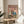 May include: Framed poster with pink background showing two ducks - one wearing chef's hat holding cooked fish, another in beanie with phone showing confused expression. Kitchen scene with cutting board, wine bottles, and prepared dishes scattered around. Natural wood frame sits on round wooden side table next to beige upholstered chair in bright room with plant and white curtains.
