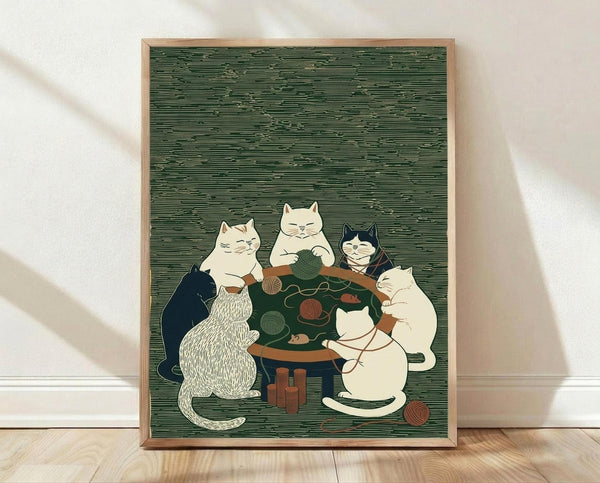 May include: Framed poster print displayed on light wooden floor against white wall with natural lighting. Green textured background artwork shows five cats gathered around wooden table playing with colorful yarn balls. Cats include white, cream, black and white, and striped varieties in whimsical illustrated style. Light wood frame surrounds the print.
