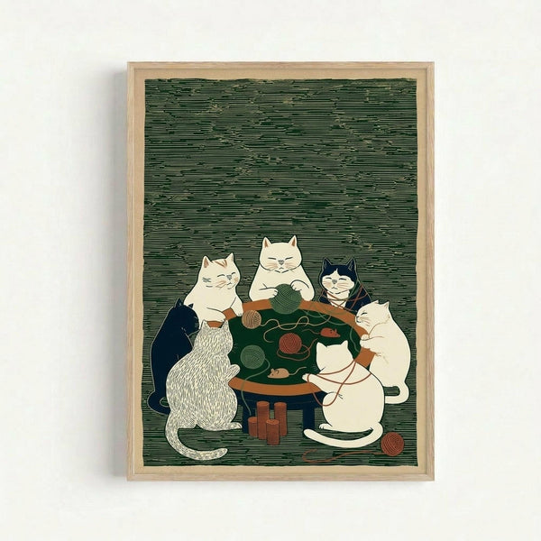 Framed artwork showing five cats gathered around a wooden table with green surface, knitting and playing with colorful yarn balls. The cats include white, black, striped, and cream colored felines in a cozy domestic scene. The illustration has a vintage woodcut style with fine line details against a textured green background. The piece is displayed in a light wooden frame on a white wall.
