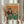 May include: Person holding framed vintage-style poster showing two anthropomorphic cats playing poker at green felt table. Male tabby cat wears sunglasses, burgundy jacket, smoking cigar. Female orange tabby wears pearl necklace, floral dress, holds martini glass. Poker chips, whiskey bottle, and playing cards scattered on table. Orange background with "CAT POKER" text in colorful letters. Light wood frame, dried pampas grass visible in bright room setting.