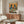 May include: A framed poster showing two anthropomorphic pigeons in vintage attire sitting at a green table with brunch items including avocado toast, pancakes, and coffee cups. The artwork features warm orange background and colorful text reading "PIGEON BRUNCH" at top. The poster is displayed above a mid-century modern walnut credenza in a minimalist living room with white walls, cream chair, glass coffee table, and white table lamp.