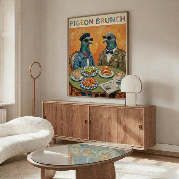 May include: A framed poster showing two anthropomorphic pigeons in vintage attire sitting at a green table with brunch items including avocado toast, pancakes, and coffee cups. The artwork features warm orange background and colorful text reading "PIGEON BRUNCH" at top. The poster is displayed above a mid-century modern walnut credenza in a minimalist living room with white walls, cream chair, glass coffee table, and white table lamp.