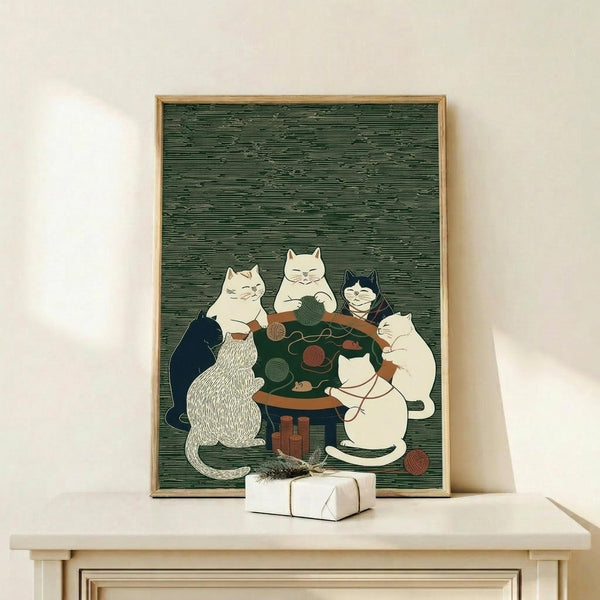 May include: Framed artwork showing five cats gathered around a small wooden table with yarn balls and knitting supplies, displayed on a light wooden shelf with a wrapped gift box. The illustration features cats in various colors - white, black, striped, and cream - against a textured green background. The print appears in a thin wooden frame leaning against a white wall.