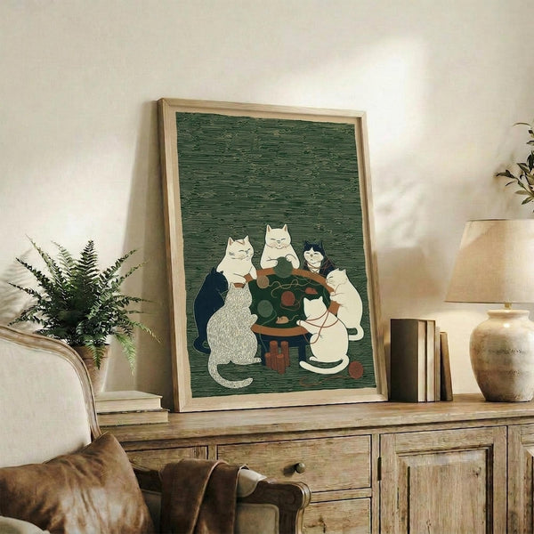 May include: Framed artwork showing several cats gathered around a wooden table with balls of yarn in various colors. The illustration has a green textured background and features white, black, and gray cats in a vintage folk art style. The piece is displayed on a rustic wooden sideboard alongside books, a ceramic vase, and a fern plant, with a beige armchair partially visible in a cozy living room setting.