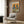May include: Framed poster with two stylized pigeons wearing vintage clothing and sunglasses sitting at a round table with brunch items including pancakes, croissants, and coffee cups against an orange background. Poster hangs on wood paneled wall above cream colored mid-century modern armchair with geometric patterned throw pillow. Modern brass floor lamp and black side table with open book complete the sophisticated living room setting.