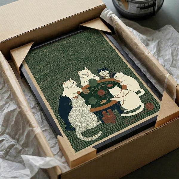 May include: Framed artwork showing six cats gathered around a green table playing with yarn balls and knitting supplies, rendered in vintage illustration style with muted green background. Print displayed in light wood frame, partially unboxed with brown cardboard packaging and white tissue paper on wooden surface.
