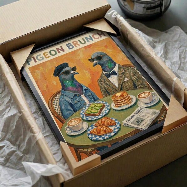 May include: Framed poster artwork showing two anthropomorphic pigeons in vintage clothing sitting at a brunch table with avocado toast, croissants, pancakes and coffee cups. Orange background with "PIGEON BRUNCH" text at top. Print displayed in black frame, partially unwrapped from cardboard packaging with protective materials on white surface.