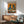 May include: Framed poster showing two pigeons in human clothing sitting at a brunch table with food and coffee, displayed above a wooden sideboard in a mid-century modern living room with gray upholstered chair, white table lamp, and natural lighting casting shadows on white walls.