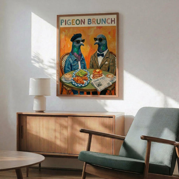 May include: Framed poster showing two pigeons in human clothing sitting at a brunch table with food and coffee, displayed above a wooden sideboard in a mid-century modern living room with gray upholstered chair, white table lamp, and natural lighting casting shadows on white walls.