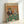 May include: Framed poster print leaning against white wall showing two anthropomorphic cats playing poker at green felt table. Left cat wears sunglasses, burgundy jacket, smoking cigar while holding cards. Right cat wears pearl necklace, patterned vest, holds martini glass. Poker chips, whiskey bottle, and fish-shaped snacks scattered on table. Four aces visible. Warm orange background with vintage illustration style in light wood frame.