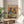 May include: Framed poster displaying two anthropomorphic pigeons in vintage clothing and sunglasses sitting at a round cafe table with brunch items including avocado toast, pancakes, croissants, and coffee cups with latte art. The illustration features vibrant orange background with colorful text reading "PIGEON BRUNCH" at top. Frame appears natural wood, positioned on light wooden side table next to white upholstered chair and potted palm plant in bright, sunlit room.