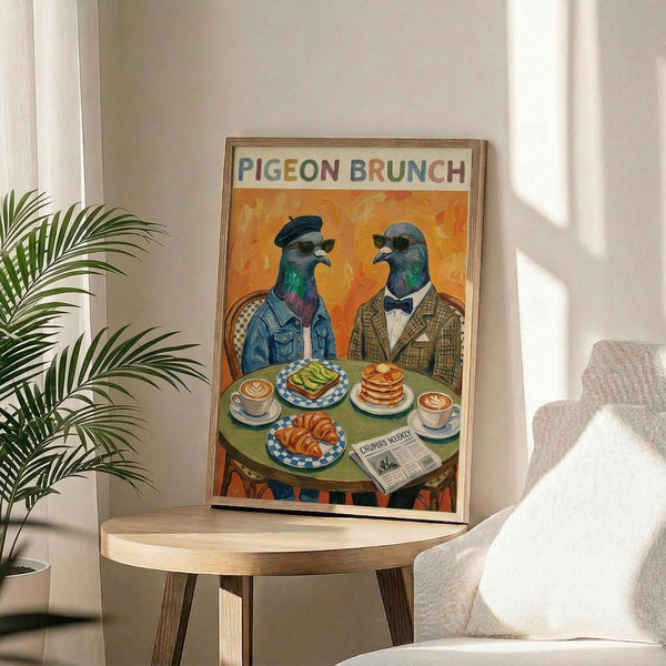 May include: Framed poster displaying two anthropomorphic pigeons in vintage clothing and sunglasses sitting at a round cafe table with brunch items including avocado toast, pancakes, croissants, and coffee cups with latte art. The illustration features vibrant orange background with colorful text reading "PIGEON BRUNCH" at top. Frame appears natural wood, positioned on light wooden side table next to white upholstered chair and potted palm plant in bright, sunlit room.