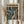 May include: Hands holding a framed anime-style poster featuring a character with dark hair in a brown coat standing in a rainy cyberpunk city street. The artwork has Japanese text at the top and shows neon signs and urban buildings in teal and orange tones. A bundle of dried wheat stalks sits nearby against soft neutral curtains in natural lighting.