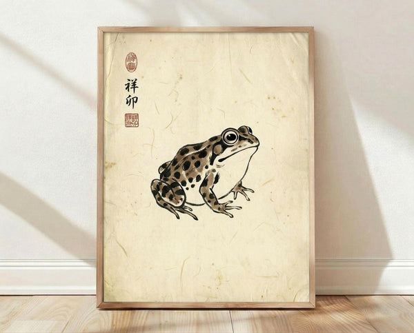May include: Framed poster print featuring a spotted frog in Japanese ink wash painting style on aged cream paper with black Chinese characters and red seal stamps, displayed in light wood frame leaning against white wall on wooden floor with soft natural lighting and shadows