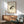 May include: Framed artwork showing a brown and black spotted frog rendered in Japanese ink wash painting style on aged cream paper with red seal stamps and black calligraphy characters. The print hangs above a wooden mid-century modern credenza with a white ceramic lamp, next to a sage green upholstered armchair with wooden frame. Warm sunlight streams across the white wall creating geometric shadows.
