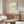 May include: Beige poster with black text arranged in a spiral pattern around a small record icon, displayed in wooden frame above mid-century modern walnut credenza. Room features cream upholstered chair, glass-top coffee table, and white table lamp in minimalist setting with neutral walls.