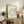May include: Framed beige poster with elegant typography arranged in spiral text pattern, displayed on rustic wooden sideboard alongside potted fern, ceramic vase, and stacked books. Warm neutral tones with natural lighting in cozy living room setting.